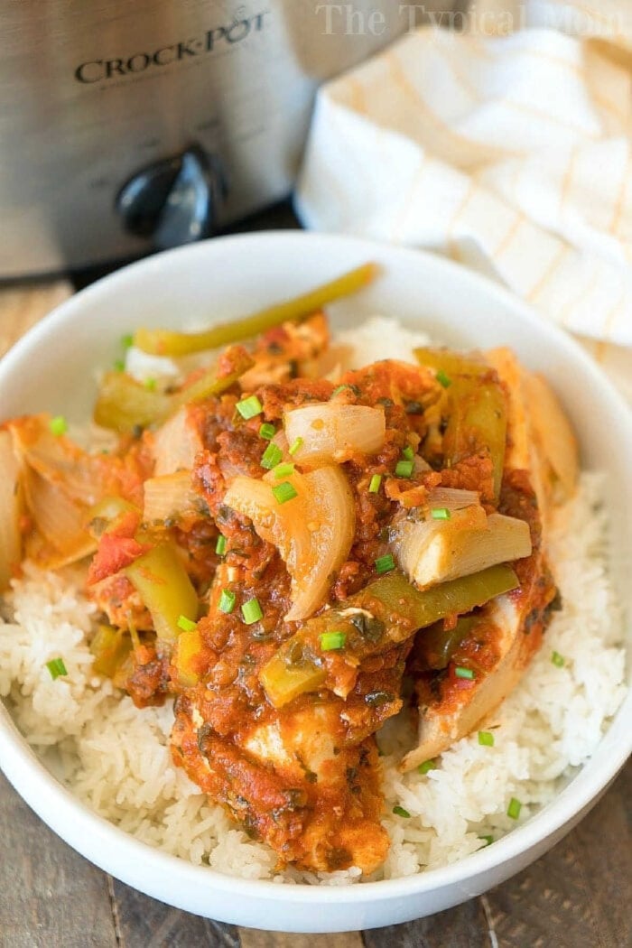 Salsa chicken crockpot recipe that only requires a few ingredients and is a healthy slow cooker dinner for sure! Shred it and make tacos or serve it on top of rice for a delicious meal packed with vegetables. If you need an easy family friendly dinner that is inexpensive but super flavorful this is it. #crockpot #slowcooker #salsachicken #chicken #healthy #salsa #crockpotrecipes #thetypicalmom A bowl of white rice topped with a saucy chicken, onion, and bell pepper dish, made using an easy salsa chicken crockpot recipe, with a slow cooker in the background.
