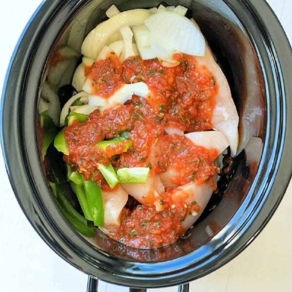 Slow cooker salsa chicken with raw chicken, sliced onions, and green peppers in a black slow cooker.