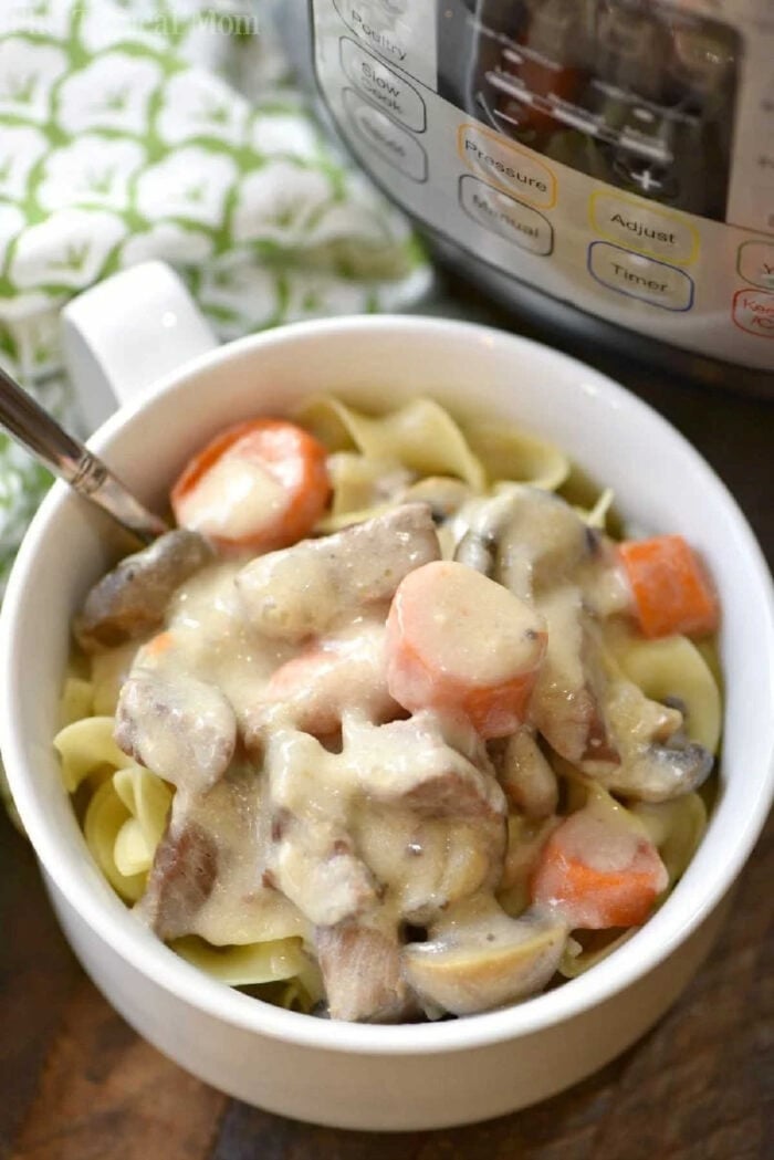 Easy Instant Pot beef stroganoff recipe that is quick and as creamy as you remember from your childhood! Total comfort food made in your pressure cooker in a fraction of the time it used to. #instantpot #pressurecooker #beef #stroganoff #instantpotrecipes #thetypicalmom A bowl of Instant Pot beef stroganoff with stew meat, carrots, and mushrooms served over noodles sits next to the pressure cooker.