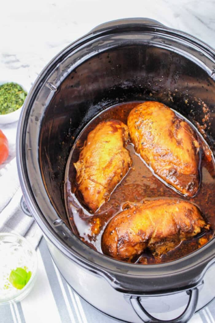 Three seasoned Crockpot Chicken Frozen Breasts cook gently in a slow cooker with savory sauce, resting on a striped cloth.