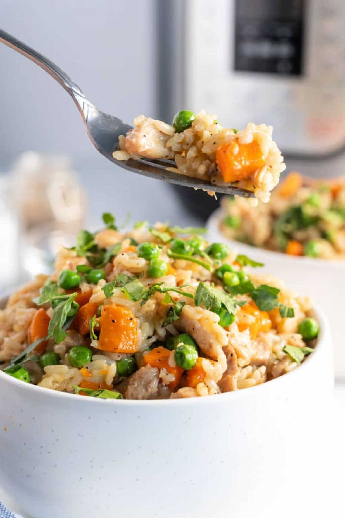 A bowl of instant pot chicken thighs and rice with peas, carrots, and herbs, as a fork lifts a delicious bite above it.