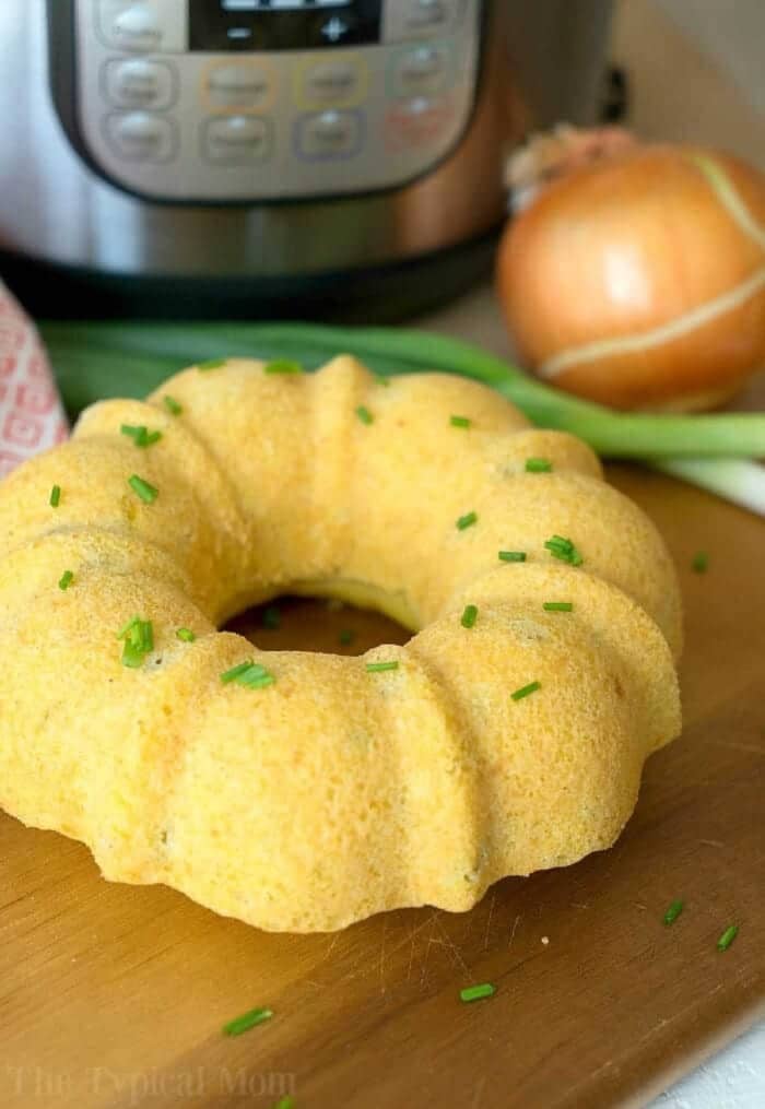 Pressure cooker cornbread is the perfect side dish for any holiday and perfect served with chili! An easy homemade cornbread recipe cooked right in your Instant Pot and comes out so pretty using this bundt pan. #pressurecooker #instantpot #cornbread #instantpotrecipes #homemade #bundtcake #sidedish #bread #thetypicalmom Bundt-shaped pressure cooker cornbread topped with chopped chives on a cutting board, with an onion and Instant Pot behind.