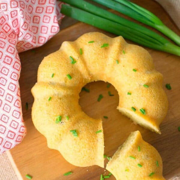 A sliced pressure cooker cornbread bundt garnished with chives sits on a wooden board, next to green onions and a red towel.