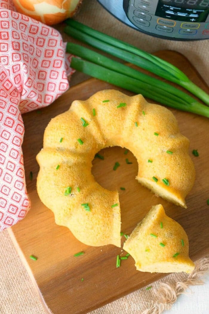 Pressure cooker cornbread is the perfect side dish for any holiday and perfect served with chili! An easy homemade cornbread recipe cooked right in your Instant Pot and comes out so pretty using this bundt pan. #pressurecooker #instantpot #cornbread #instantpotrecipes #homemade #bundtcake #sidedish #bread #thetypicalmom A sliced pressure cooker cornbread bundt garnished with chives sits on a wooden board, next to green onions and a red towel.