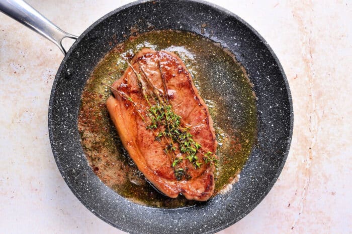 A perfectly cooked Sous Vide Frozen Steak with herbs and sauce in a black frying pan on a light surface.