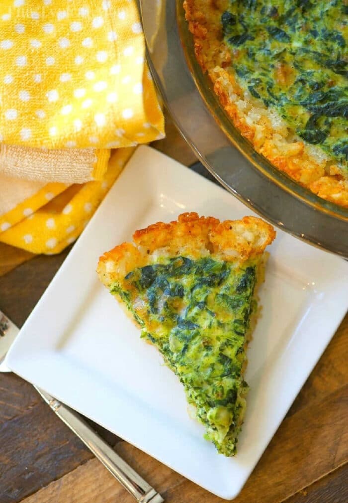 Tater tot crusted spinach quiche! It's perfect for breakfast or brunch with a crunchy tater tot pie crust and warm egg filling. #tatertots #quiche #tatertot #pie #spiach #cheese #breakfast A slice of tater tot spinach quiche with a hash brown crust sits on a white plate, next to a yellow polka dot napkin.