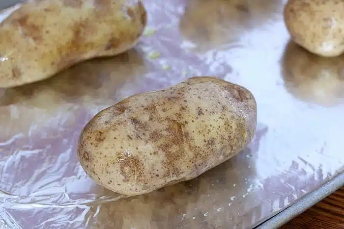 A large, raw potato coated in oil rests on a foil-lined baking sheet, perfect for prepping Make Ahead Twice Baked Potatoes. Another potato peeks from the background, waiting its turn.