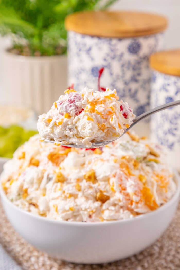 Easy ambrosia recipe for you that just takes a few ingredients and is really yummy. Dessert side dish with fruit and marshmallows our whole family loves. #ambrosia #salad #fruit #sidedish #marshmallow A spoonful of creamy fruit salad held above a bowl filled with the same salad, showcasing this easy ambrosia recipe.