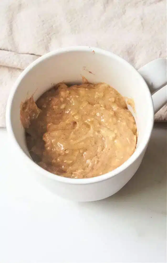 A white mug holds a thick mixture of mashed bananas and peanut butter, reminiscent of banana bread in a mug, sitting on a white surface with a beige cloth in the background.