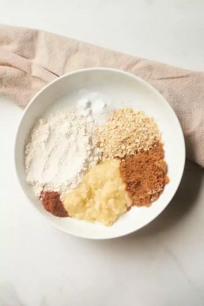 A white bowl holds the makings of banana bread in a mug: flour, oats, brown sugar, mashed banana, cinnamon, and a dash of baking soda. A beige cloth subtly peeks into view on the pristine white surface.