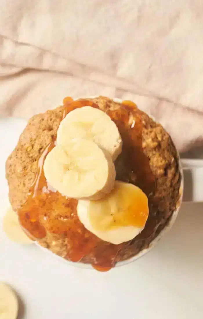 A banana bread mug cake topped with sliced bananas and drizzled with syrup is placed against a soft pink background. Some banana slices are also visible on the surface near the mug.