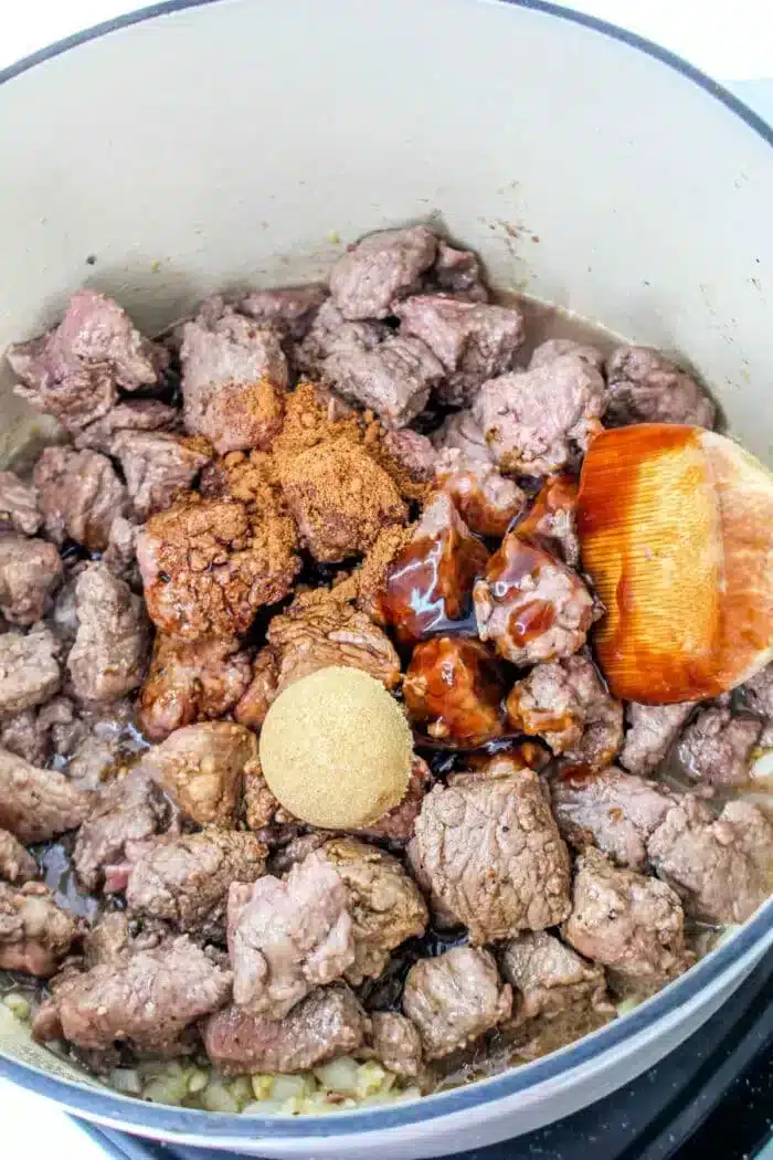 A pot containing braised beef tips cooked with spices, brown sugar, and a savory sauce. A wooden spoon is partially visible, resting on the edge of the pot.