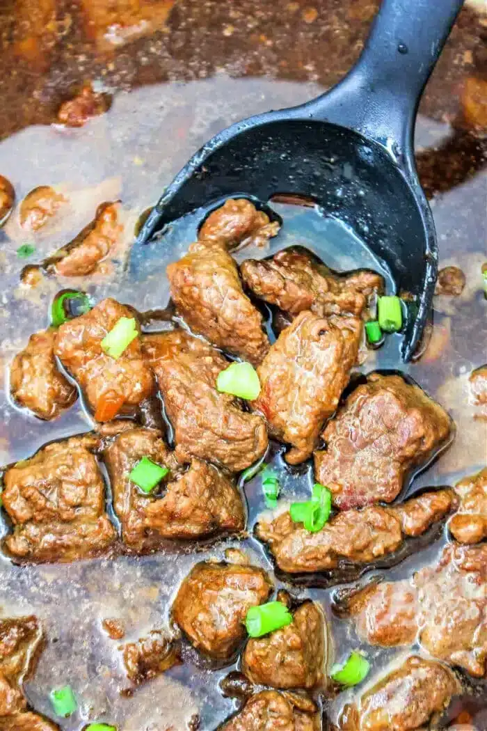 A ladle filled with succulent Braised Beef Tips in a rich brown sauce, garnished with chopped green onions. The tender beef pieces are submerged in the savory sauce, and the ladle is black.