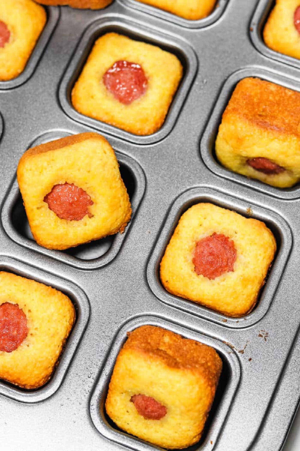 Mini square Corn Dog Bites in a metal baking tray, each with a piece of hot dog in the center.