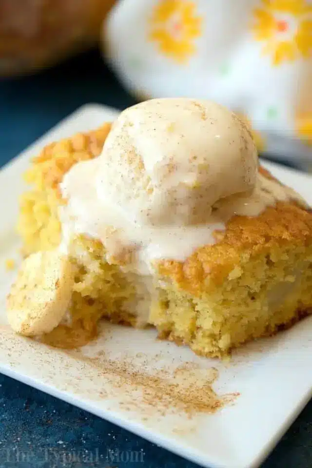 A slice of cake, made using clever box cake mix hacks, is topped with a scoop of vanilla ice cream on a white square plate. The cake boasts a light golden color, with a small cookie on the side. Cinnamon graces the top and surrounds the dessert, set against a backdrop of patterned cloth.