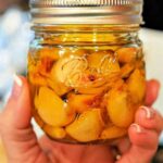 A hand tenderly holds a jar of preserved garlic cloves in oil, capturing the essence of how to make garlic confit.