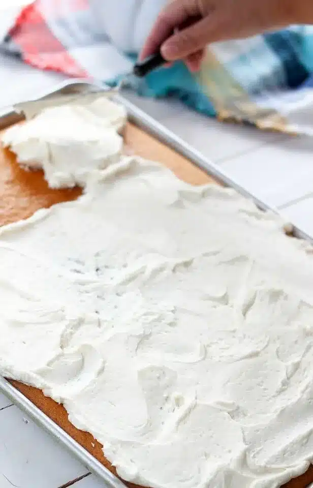 A hand is expertly using a spatula to spread white frosting over a large sheet cake on a metal baking tray, showcasing one of the best box cake mix hacks. A colorful cloth peeks in the background, adding a touch of whimsy to this delicious creation.