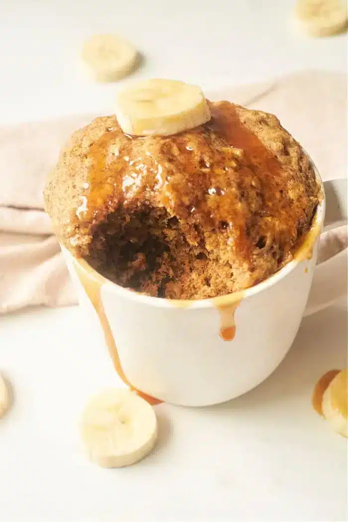 A Banana Bread in a Mug creation, topped with a banana slice and drizzled with syrup. The cake has a bite taken out of it, revealing its moist texture. Additional banana slices are artfully scattered around the mug on a light surface, enhancing its delicious appeal.