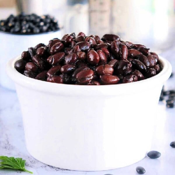 A white bowl filled with perfectly tender black beans cooked in a pressure cooker rests on a marble surface, with dried beans scattered in the background.