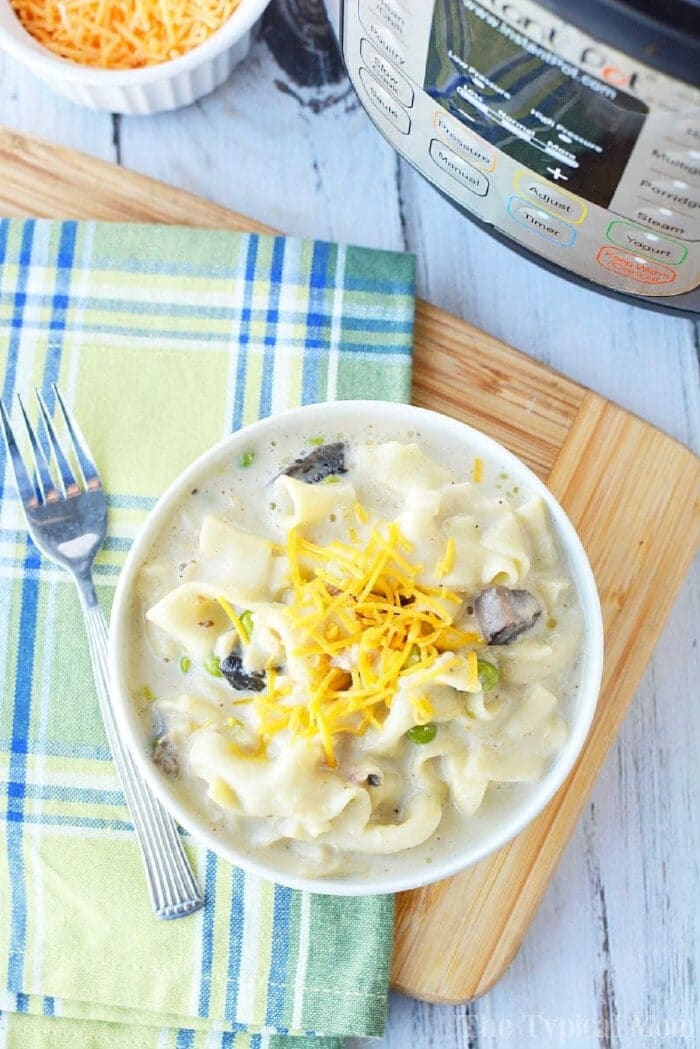Instant Pot tuna noodle casserole will bring you back to your childhood for sure. The comfort food you love can now be made in your Instant Pot for dinner in no time at all and is loaded with all the ingredients you once made in your oven. #instantpot #pressurecooker #tunanoodlecasserole #casserole #tuna #tunacasserole #instantpotrecipes #thetypicalmom A bowl of creamy noodle soup inspired by pressure cooker tuna noodle casserole, topped with shredded cheese, sits next to a fork and an Instant Pot.