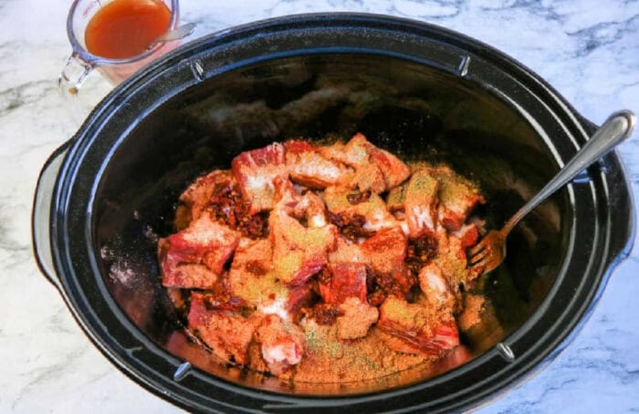 Raw pork ribs with spices in a slow cooker, reminiscent of slow cooker beef barbacoa, with a fork and a cup of sauce nearby on a marble surface.