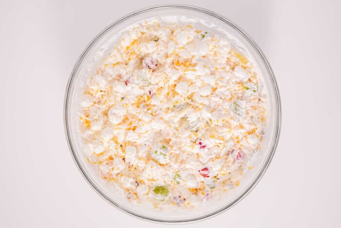 Easy ambrosia recipe for you that just takes a few ingredients and is really yummy. Dessert side dish with fruit and marshmallows our whole family loves. #ambrosia #salad #fruit #sidedish #marshmallow A glass bowl filled with a creamy salad mixture containing marshmallows, fruit, and shredded cheese—an easy ambrosia recipe perfect for any occasion.
