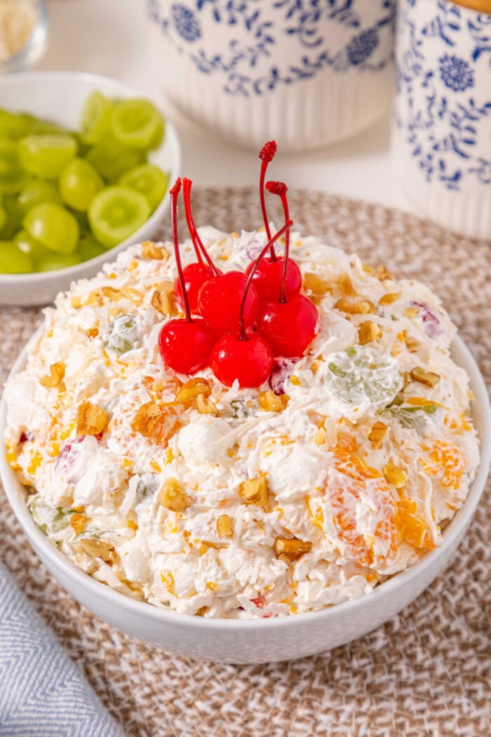 A bowl of fruit salad topped with five maraschino cherries, with grapes in the background—just like an easy ambrosia recipe served fresh.