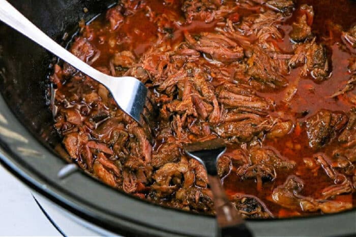 Slow-cooked shredded beef barbacoa in rich sauce, with two forks pulling apart the tender meat in a black slow cooker.