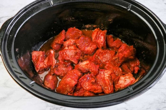 Chunks of raw beef coated in marinade inside a black slow cooker, ready to become tender slow cooker beef barbacoa on a marble surface.