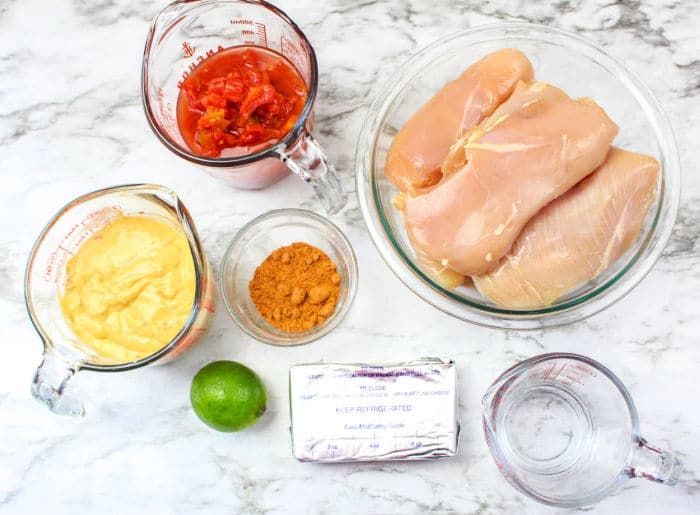 Raw chicken breasts, tomatoes, soup, taco seasoning, cream cheese, lime, and water on a marble counter—everything you need for delicious Pressure Cooker Chicken Tacos.