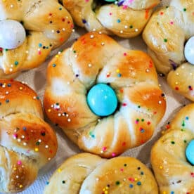 Sweet bread rolls twisted into rings, these delightful easter egg bread treats are topped with colorful eggs and rainbow sprinkles on a white background.