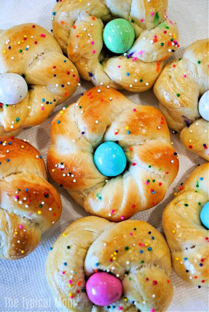 Sweet bread rolls twisted into rings, these delightful easter egg bread treats are topped with colorful eggs and rainbow sprinkles on a white background.