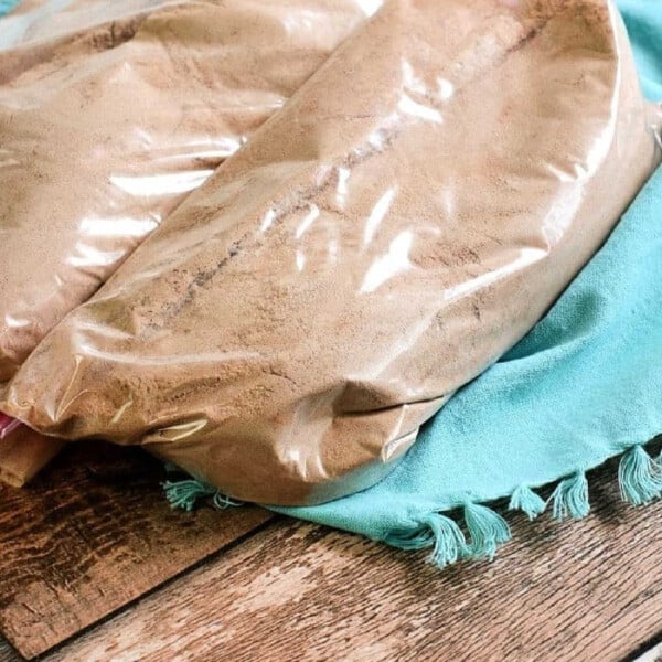 Two clear plastic bags filled with brown powder, resembling a homemade cake mix, rest on a blue fringed cloth on a wooden surface.