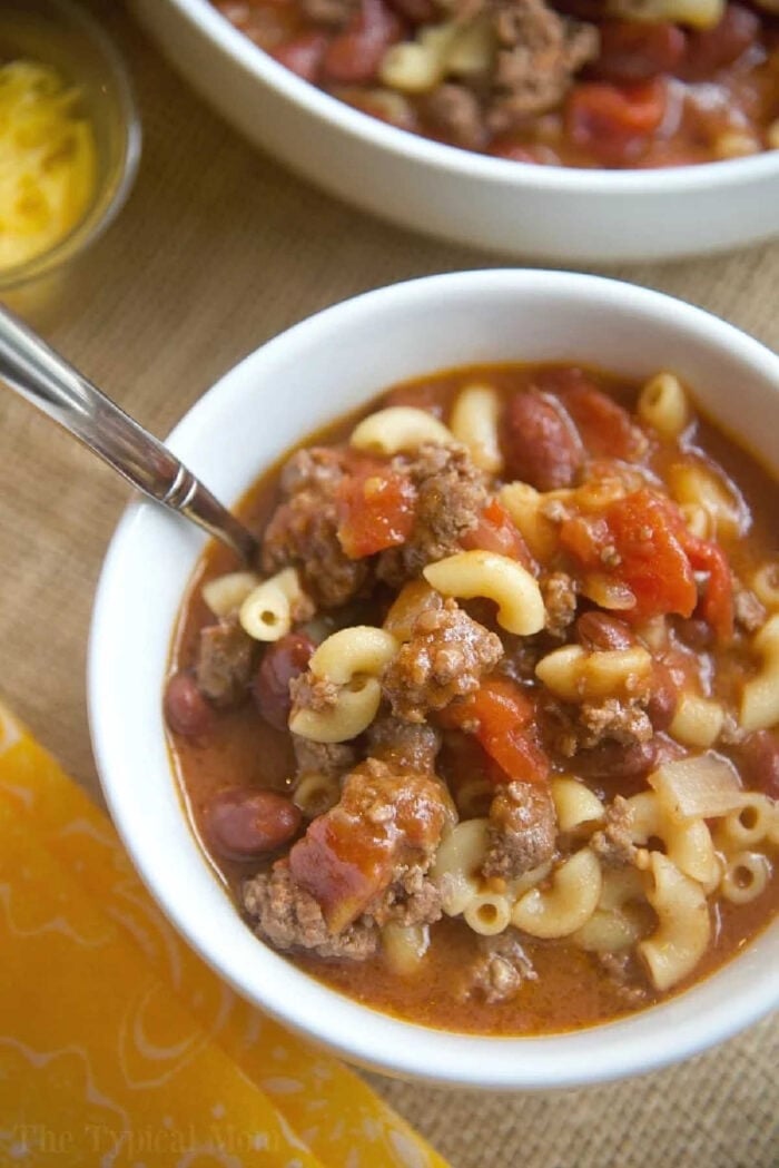 Instant Pot chili mac recipe that takes 5 minutes to cook and you and your kids will love! A classic old fashioned goulash dinner packed with flavor. #instantpot #chilimac #chili #macaroni #goulash #pressurecooker #groundbeef #instantpotrecipes #thetypicalmom #ninjafoodi #ninjafoodirecipes #crockpotexpressrecipes A bowl of goulash featuring macaroni, ground beef, tomatoes, and beans, with a spoon in the bowl—perfect for those craving an easy instant pot chili mac recipe.