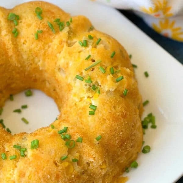 Golden cornbread ring on a white plate, garnished with chopped chives, reminiscent of an instant pot corn casserole recipe, with a floral napkin nearby.