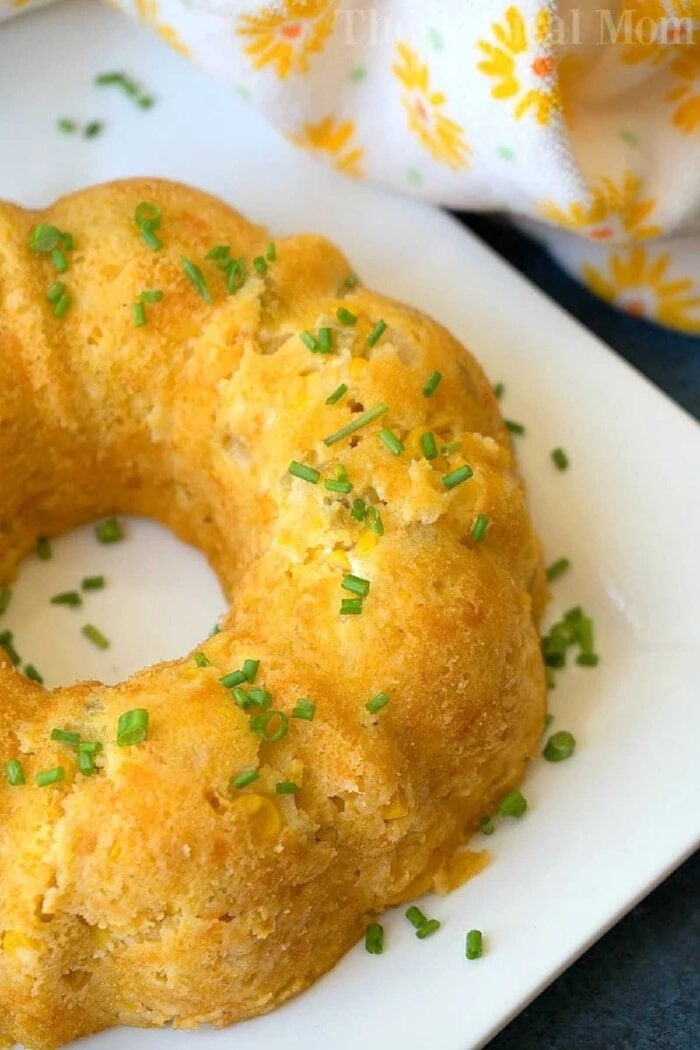 This Instant Pot corn casserole recipe is a must during the holidays. Cheesy creamed corn side dish in your pressure cooker for Thanksgiving is a must! #corncasserole #instantpot #instantpotrecipes #thetypicalmom #casserole #thanksgiving #pressurecooker #corn Golden cornbread ring on a white plate, garnished with chopped chives, reminiscent of an instant pot corn casserole recipe, with a floral napkin nearby.