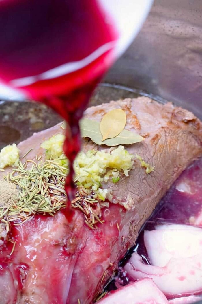 Red wine being poured over a seasoned pressure cooker roast beef with bay leaves, herbs, and garlic in a pot.