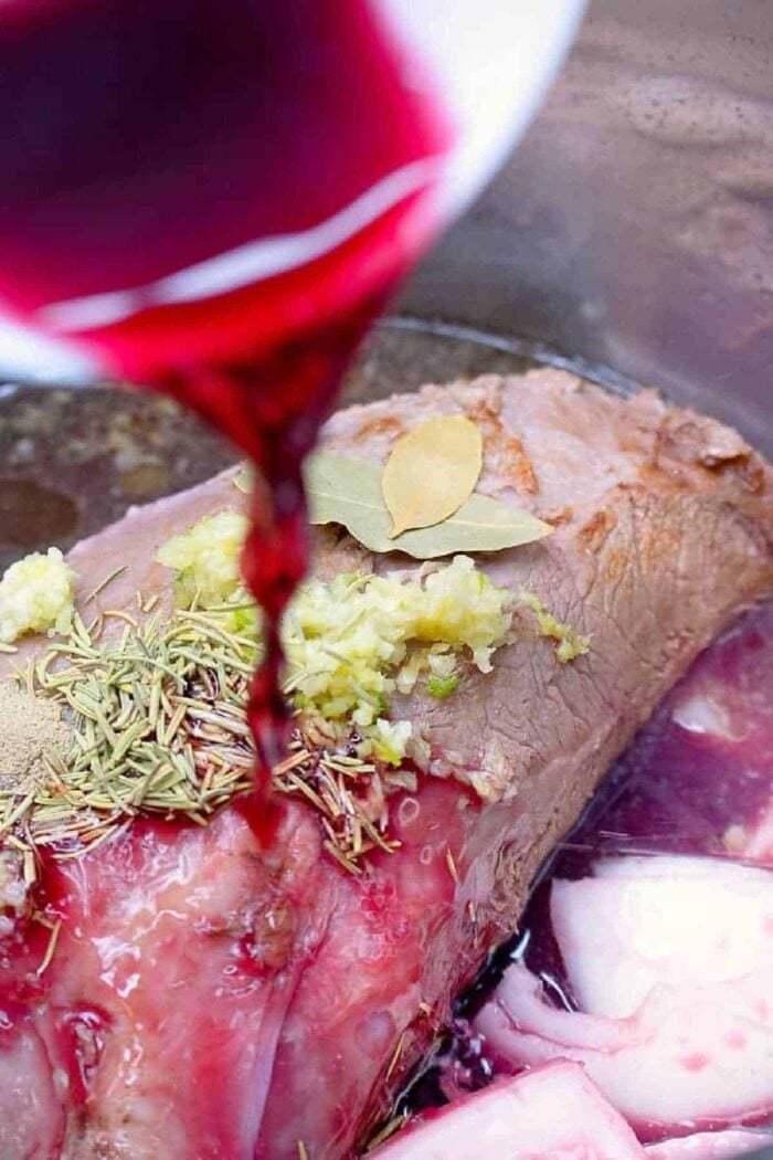 Pressure cooker roast beef is tender and full of flavor without ever having to turn on your oven! Sliced or shredded beef Instant Pot or Ninja Foodi style. #instantpotrecipes #ninjafoodirecipes #roastbeef #pressurecooker #thetypicalmom #instantpot #ninjafoodi #crockpotexpress Red wine being poured over a pressure cooker roast beef seasoned with herbs, garlic, and a bay leaf.