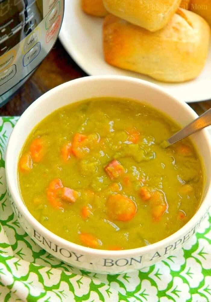 Instant Pot split pea soup recipe with ham is so easy to make and my kids love it! Throw it in your pressure cooker for 17 minutes and it's the perfect healthy soup made with dried peas that require no soaking beforehand. Fresh homemade split pea soup in less than 20 minutes! #instantpotrecipes #instantpot #pressurecooker #splitpeasoup #soup #homemade #thetypicalmom A bowl of Instant Pot Split Pea Soup Recipe with diced ham and carrots, served alongside a plate of bread rolls.