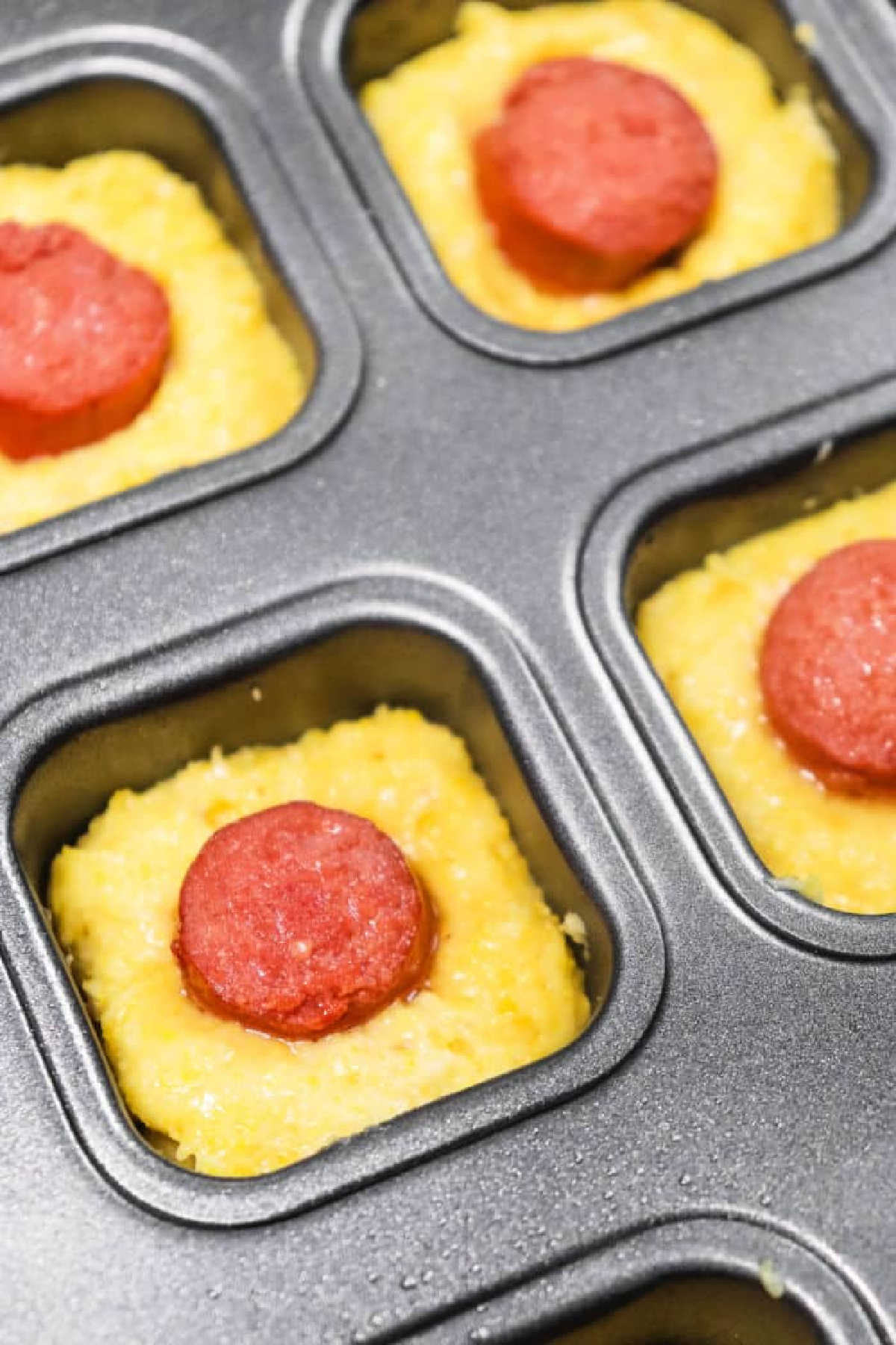 Unbaked Corn Dog Bites cornbread batter in a muffin tin, each topped with a slice of pepperoni.