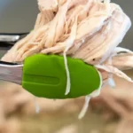 A close-up of Ninja Foodi shredded chicken caught in green kitchen tongs above a pot. The background reveals more tender, perfectly shredded chicken waiting to be served.