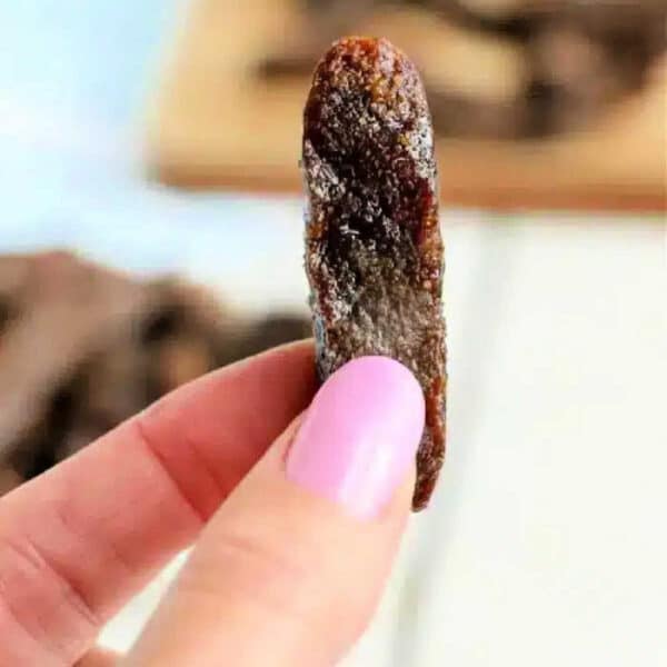 A hand with pink nail polish holds a single dried tamarind piece close to the camera, reminiscent of air fryer beef jerky in its texture and appearance.