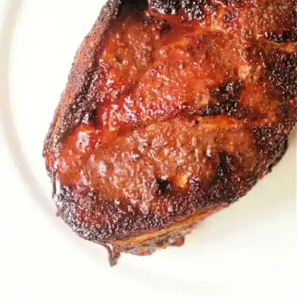 Close-up of a cooked, seasoned piece of air fryer pork roast with a charred exterior, displayed on a white plate. The meat has a rich brown color with a slightly crispy texture on the surface.