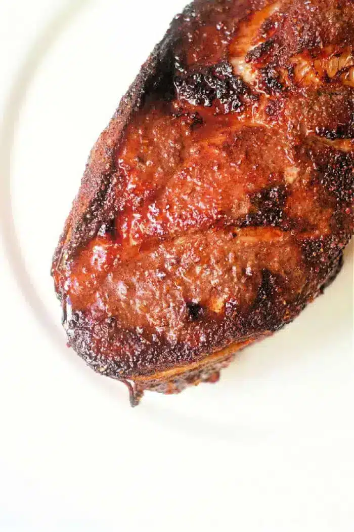 Close-up of a cooked, seasoned piece of air fryer pork roast with a charred exterior, displayed on a white plate. The meat has a rich brown color with a slightly crispy texture on the surface.