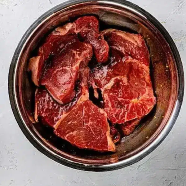 A metal bowl contains several pieces of red meat marinating in an Asian steak marinade, all placed on a gray, textured surface.