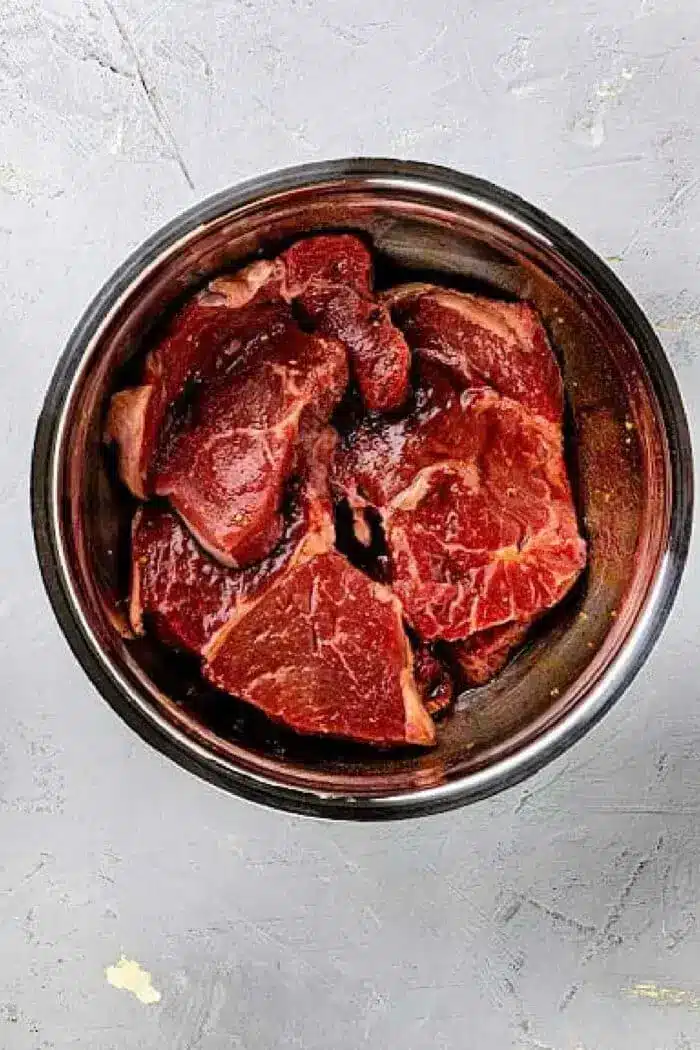 A metal bowl contains several pieces of red meat marinating in an Asian steak marinade, all placed on a gray, textured surface.