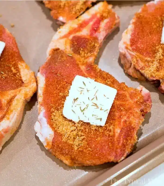 The best baked pork chops are seasoned with spices and carefully arranged on a baking tray. Each succulent chop boasts a pat of butter on top, enhanced with a sprinkle of dried herbs for perfect flavor.