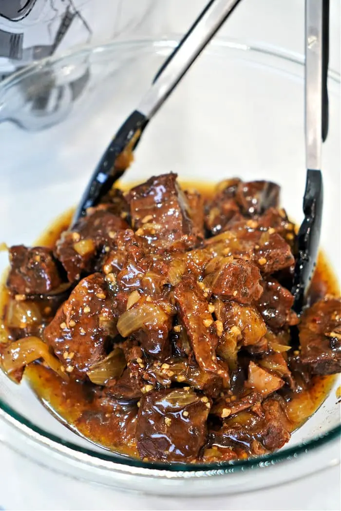 A glass bowl brims with succulent beef arm roast pieces in a thick, rich sauce. The dish is elegantly garnished with onion slices and sesame seeds, with a pair of black-handled tongs resting on the edge—perfect for savoring this delightful recipe.