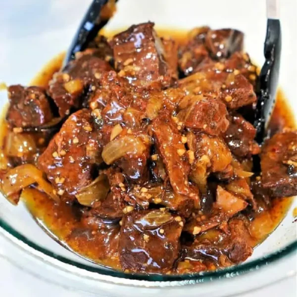 A glass bowl filled with beef stew, showcasing a classic beef arm roast recipe with chunks of beef and onions in a rich, dark sauce. Black tongs rest in the bowl, and the stew is visibly seasoned with fresh herbs and spices.