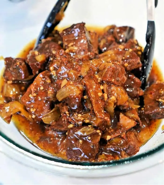 A glass bowl filled with beef stew, showcasing a classic beef arm roast recipe with chunks of beef and onions in a rich, dark sauce. Black tongs rest in the bowl, and the stew is visibly seasoned with fresh herbs and spices.